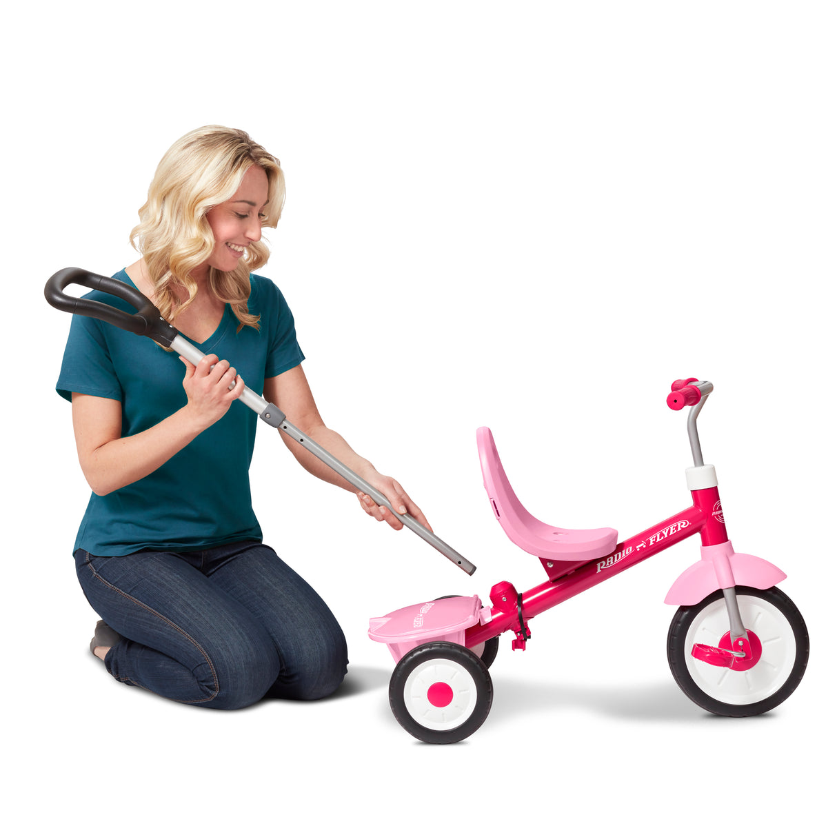 A woman kneels beside a pink Radio Flyer tricycle, holding a long-handled tool that appears to be used for adjusting the tricycle's handlebar height. The tricycle has a bright pink frame, a matching pink seat, and white wheels with pink centers.