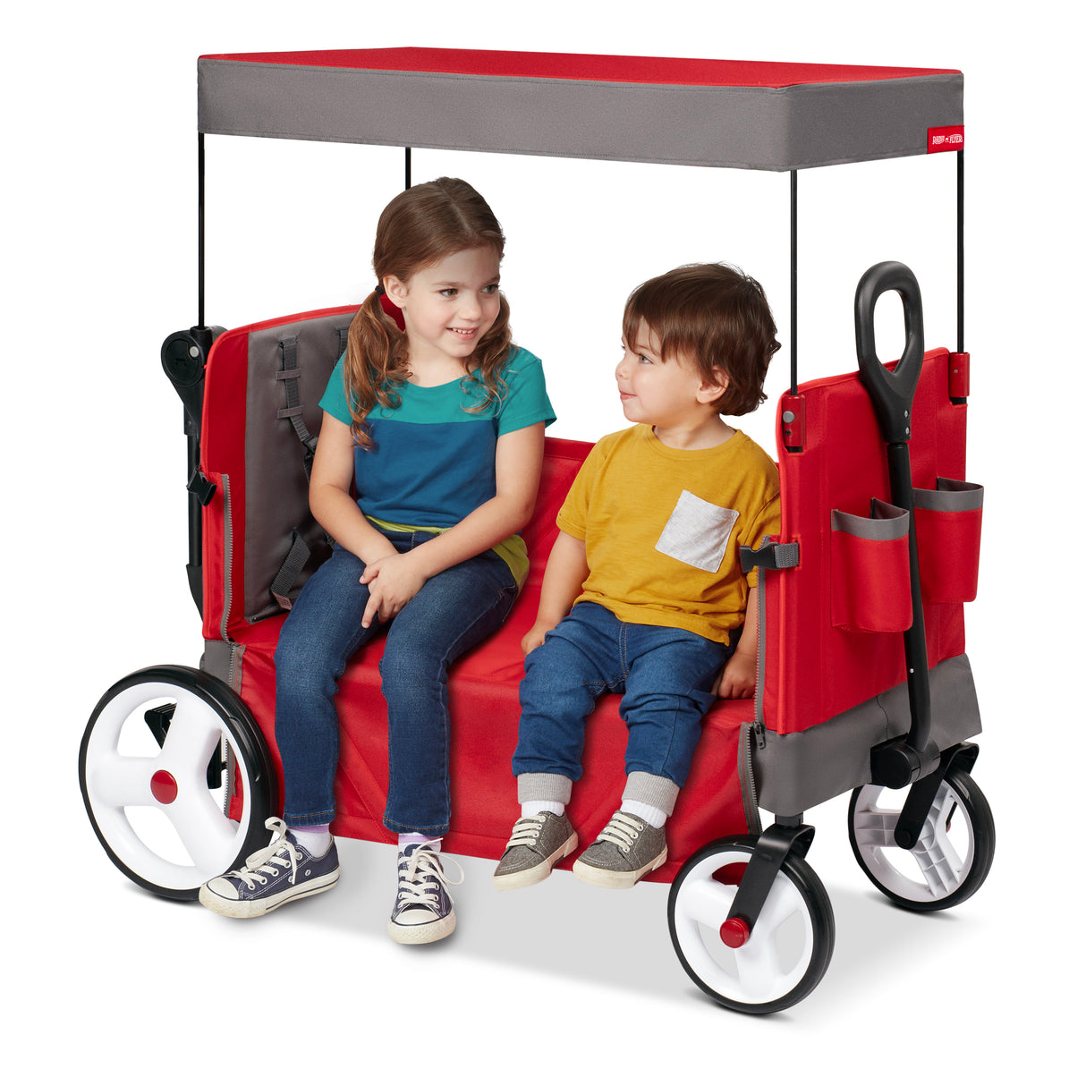 A red Radio Flyer wagon with a canopy, shown with two children sitting inside it in bench mode.