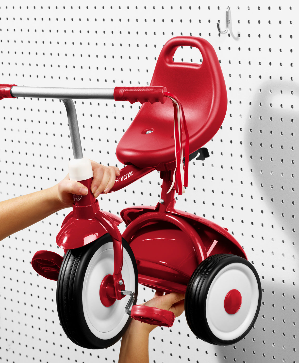 Folded red tricycle being held up and hung on a white pegboard
