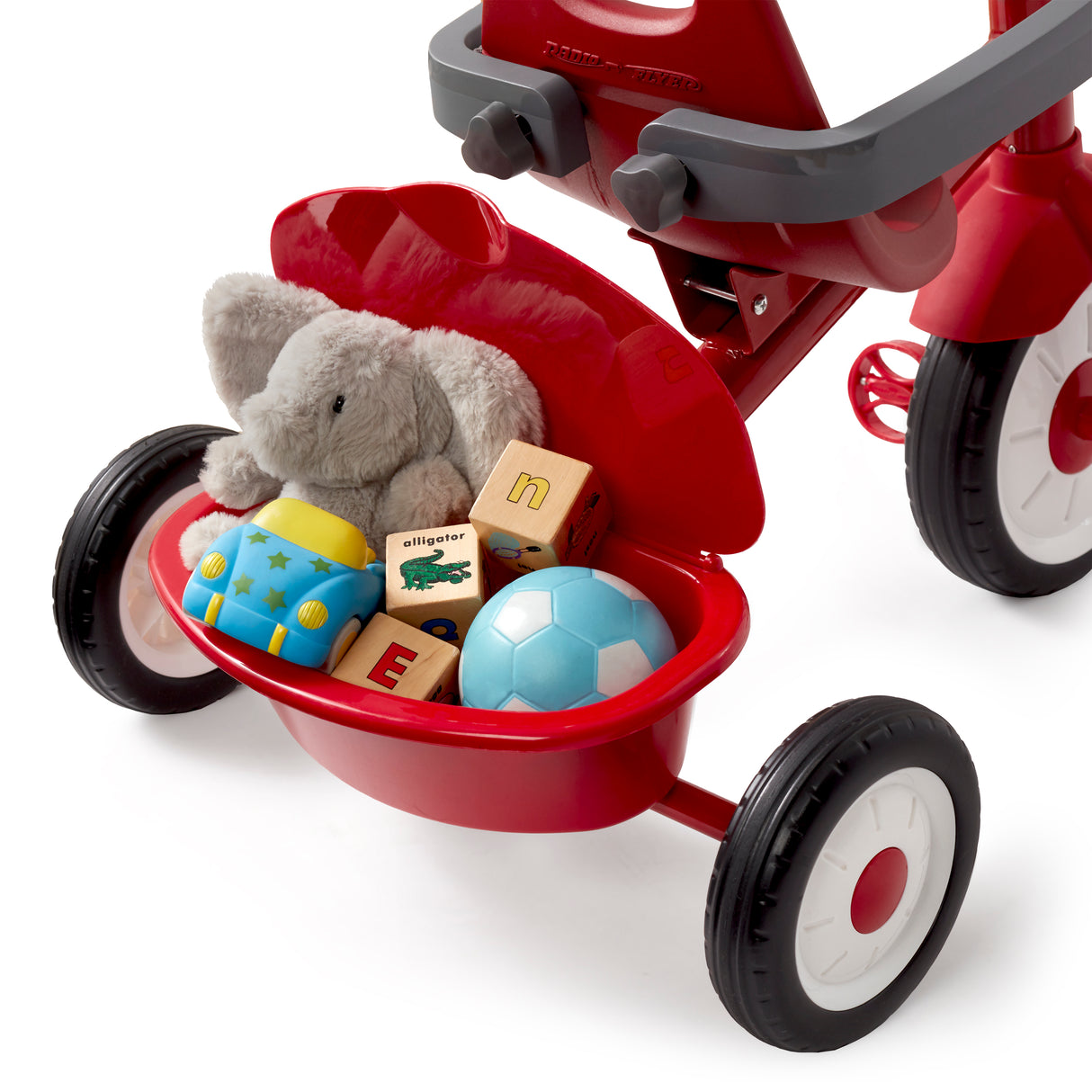 A red tricycle with a storage bin on the back, shown on a white background. The basket is filled with toys, including a gray plush elephant, wooden blocks, a blue toy, and a soccer ball.