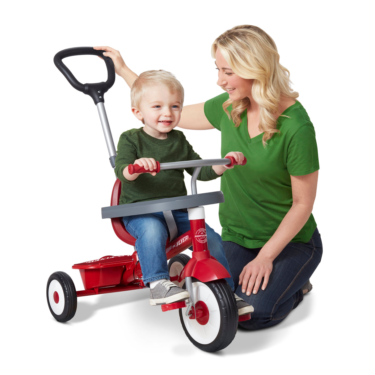 A woman in a green shirt kneels next to a young child on a red Radio Flyer tricycle with a seat and push handle, set against a solid white background.