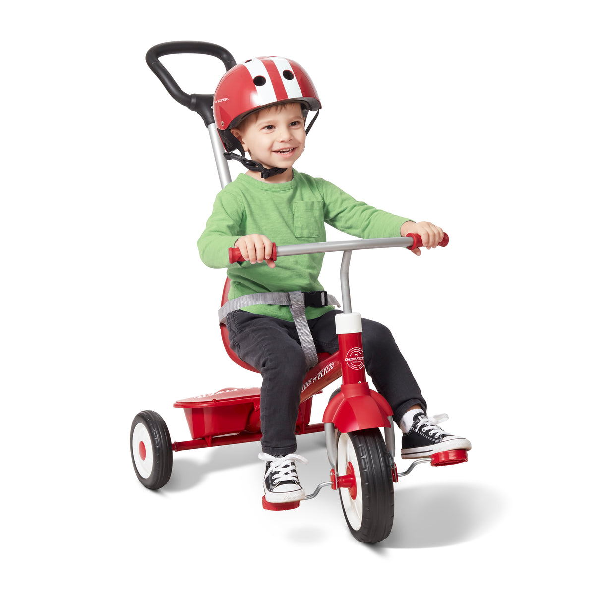 A young child wearing a red Radio Flyer helmet, green shirt, and dark pants sits on a red push-tricycle with a seat and handlebar, photographed on a clean white studio background.