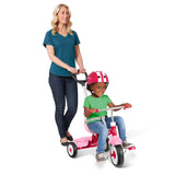 A woman pushes a pink children's tricycle with a seat and handlebar while a young child rides it, wearing a pink and white helmet, against a clean white background.