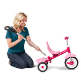 A woman kneels beside a pink Radio Flyer tricycle, holding a long-handled tool that appears to be used for adjusting the tricycle's handlebar height. The tricycle has a bright pink frame, a matching pink seat, and white wheels with pink centers.