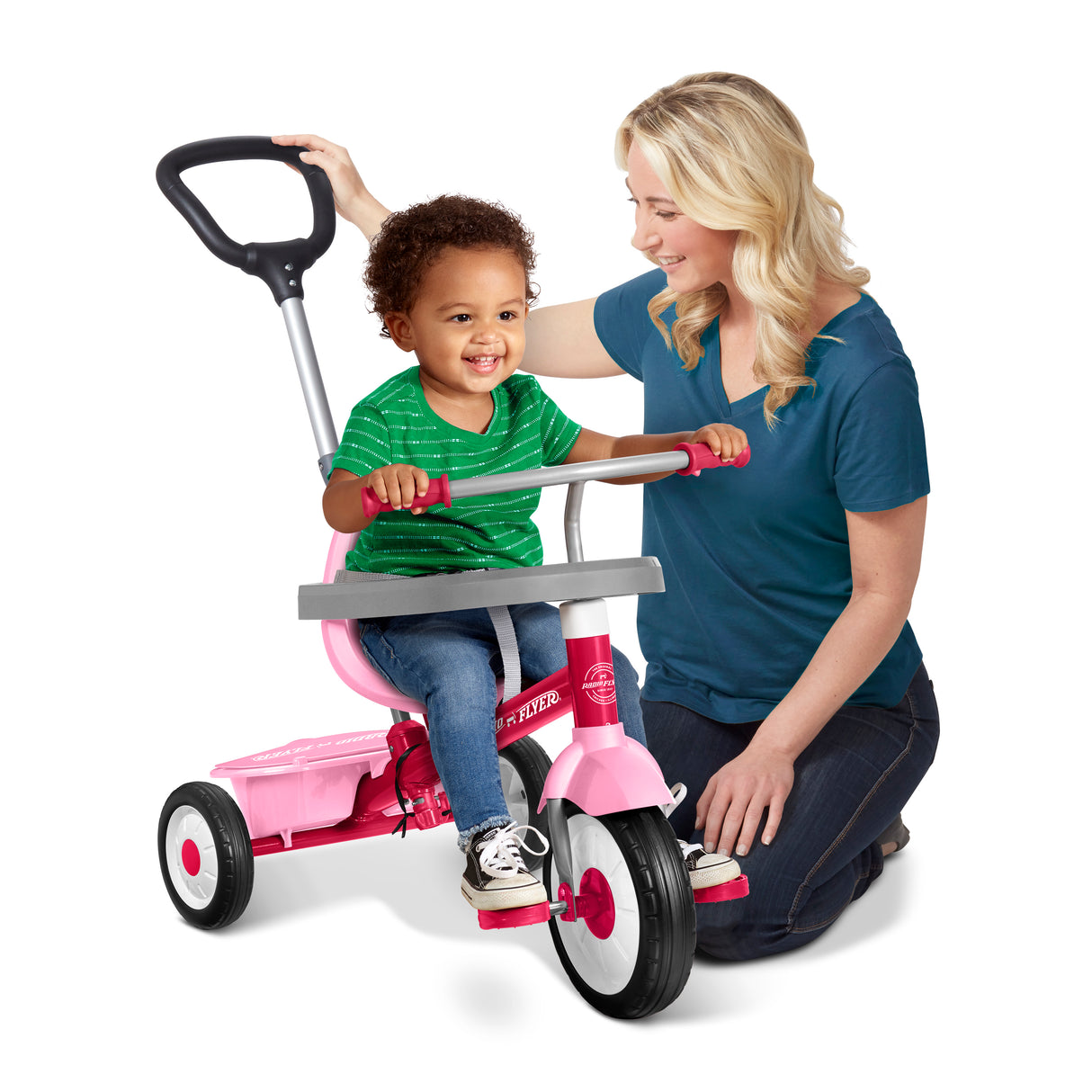 A woman kneels beside a young child sitting on a pink tricycle with a handlebar push seat, photographed on a clean white studio background.