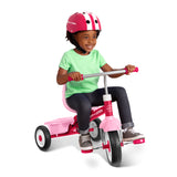 A young child riding a pink and silver tricycle with a seat and handlebars, wearing a pink helmet, green shirt, and dark pants, photographed on a clean white background.