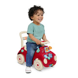 A smiling toddler with curly dark hair sits on a red and cream push toy car with colorful sensory details like gears and flipping mirrors