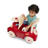 A toddler with curly dark hair opens the cream colored seat and is pulling out a small soccer ball, showcasing the under seat storage of the red ride-on push car