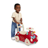 A toddler boy pushing a red and cream plastic car with a steering wheel and colorful gear details