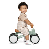 A toddler sitting on a Radio Flyer balance bike, wearing beige and white overall shorts and white sneakers, photographed against a solid white background.